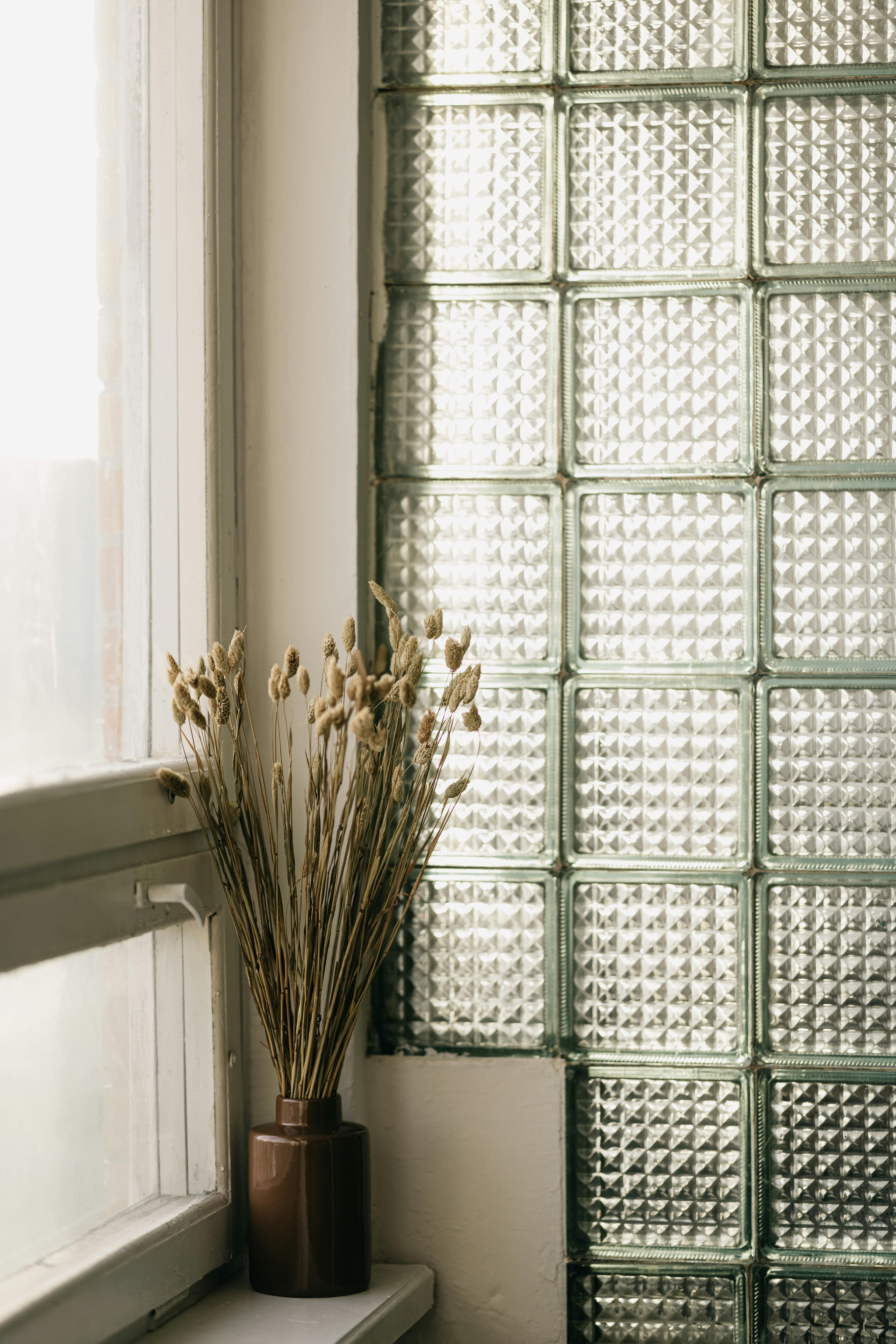 A minimalist interior featuring a vase of dried flowers on a windowsill against a textured glass wall.