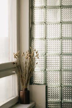 A minimalist interior featuring a vase of dried flowers on a windowsill against a textured glass wall.