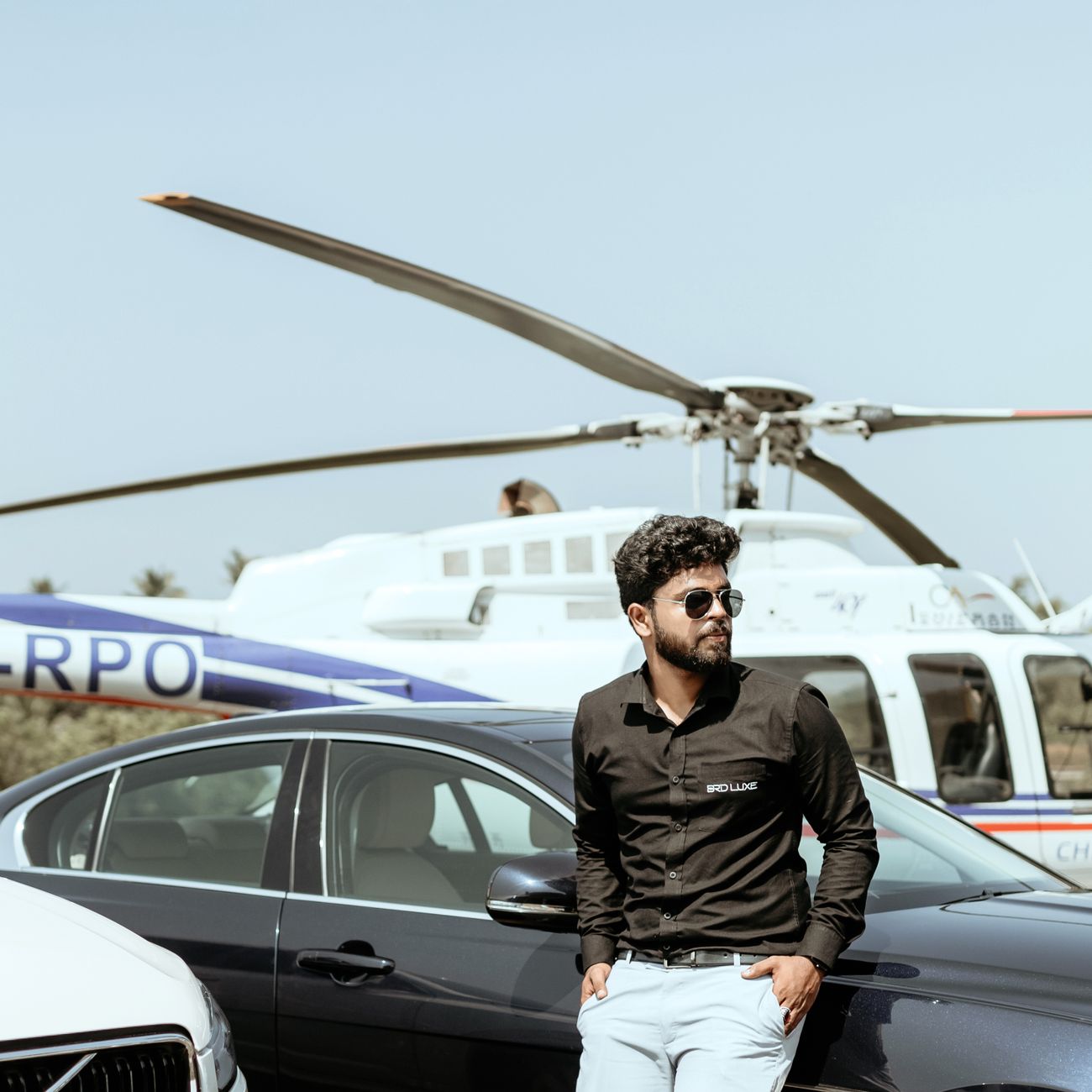Stylish man stands confidently by car and helicopter, exuding luxury lifestyle.