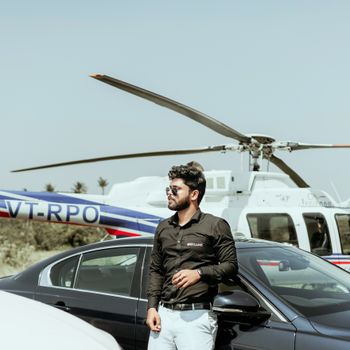 Stylish man with sunglasses stands by a car and helicopter outdoors on a sunny day.