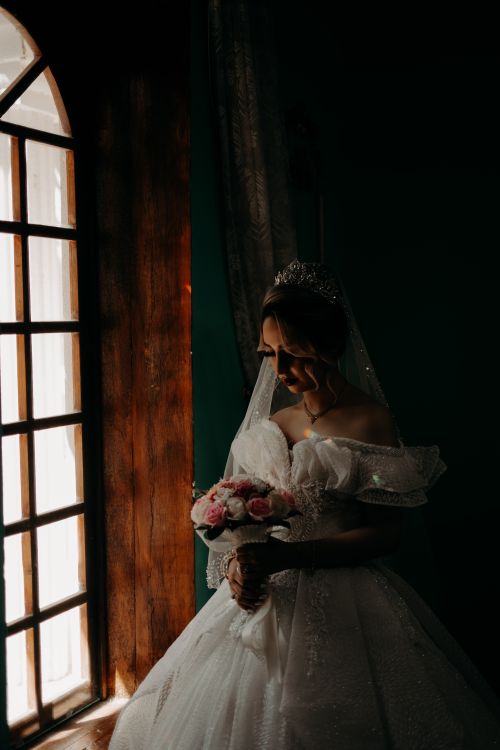 Free A beautiful bride in a wedding dress holds a bouquet while standing by a window in Tabriz, Iran. Stock Photo