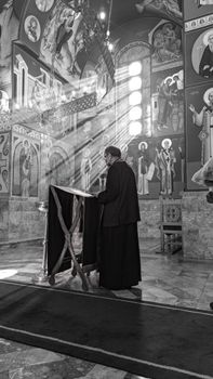 Black and white photo of a priest leading a service inside a beautifully decorated cathedral with sunlight streaming in.
