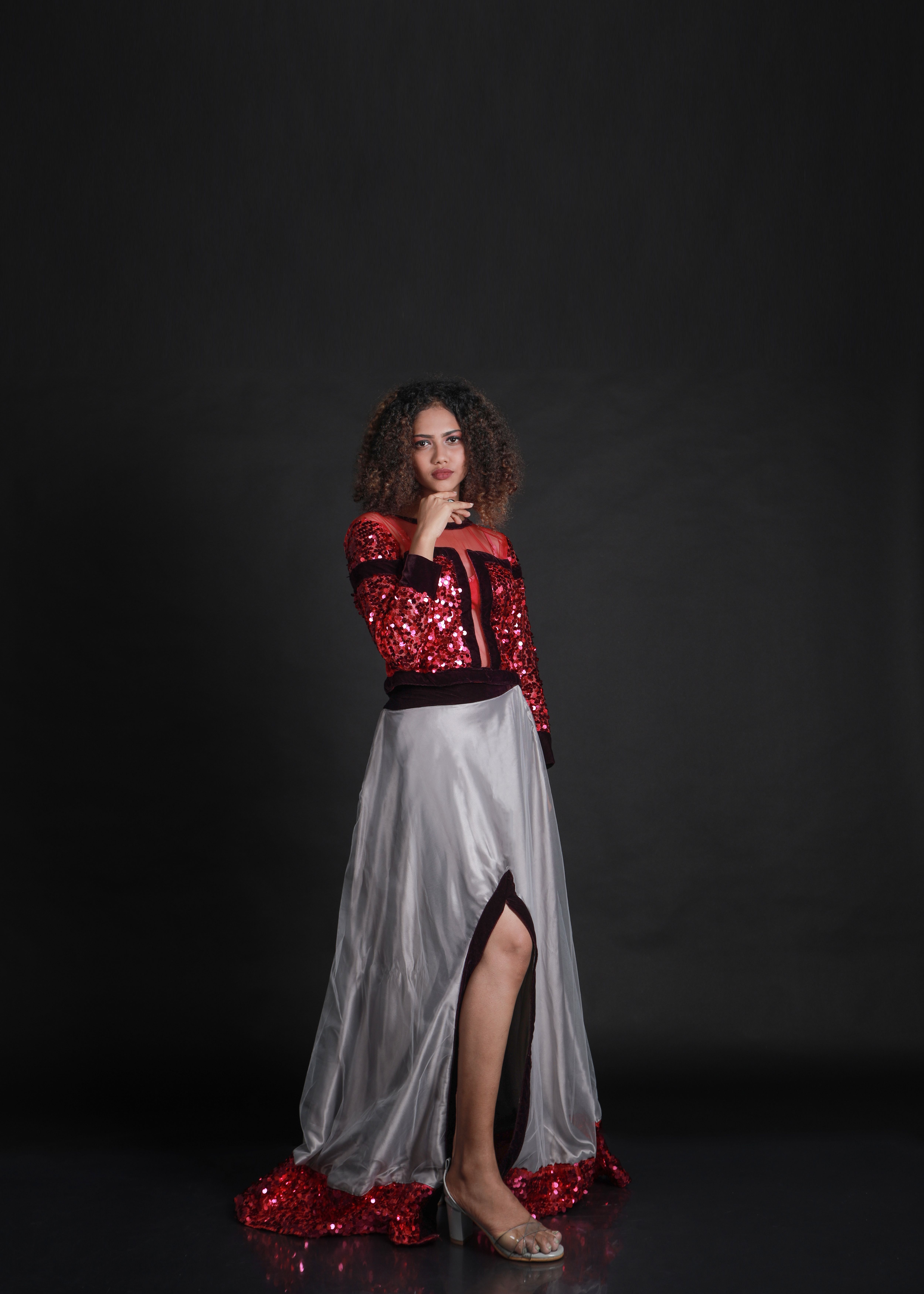 Portrait of a fashion model in a red sequined top and flowing white skirt posing against a dark studio backdrop.