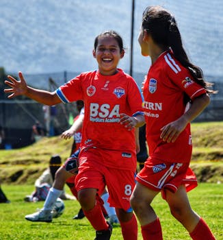 Two young girls in red uniforms joyfully playing soccer on a sunny day outdoors.