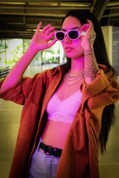 Young woman in sunglasses posing with trendy outfit under vivid purple lighting indoors.