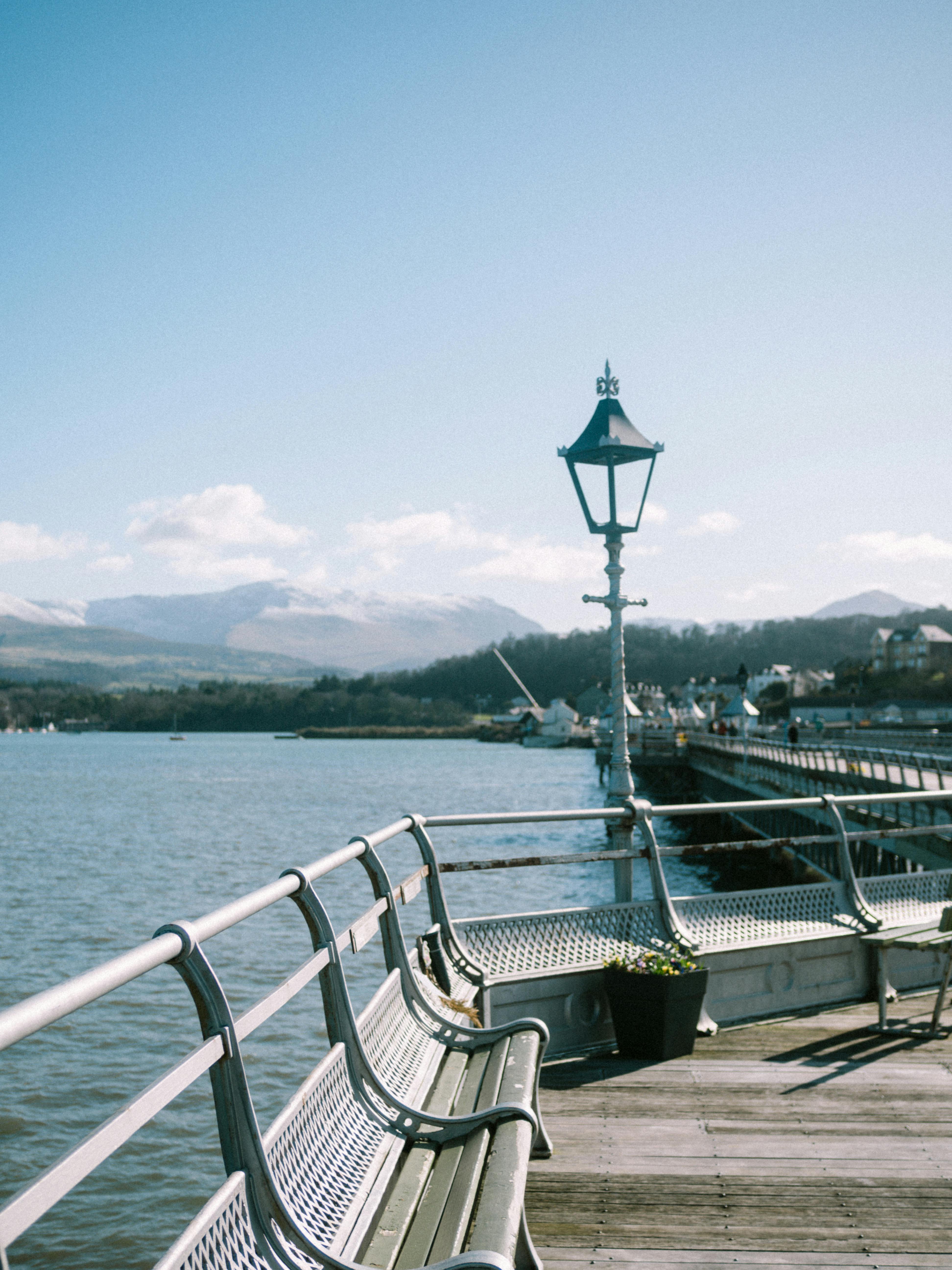A peaceful view of Bangor Pier with benches facing the Menai Strait, under a clear blue sky.