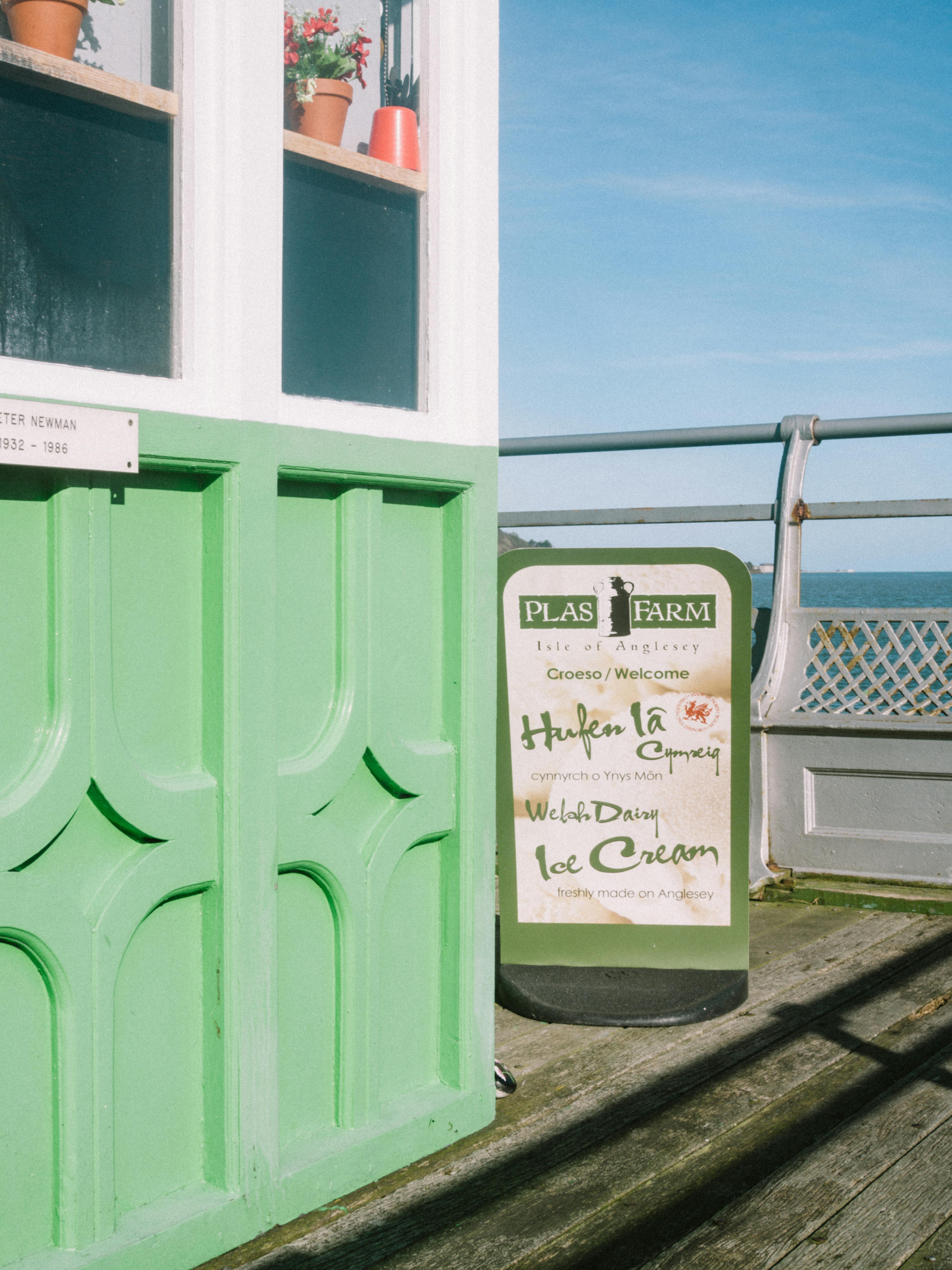 Entrance to a Welsh ice cream shop with a bright green door by the seaside pier, sunny day.