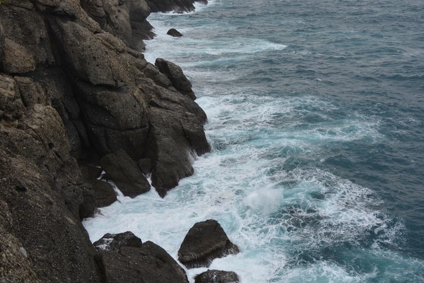 A steep rocky headland dropping into grey-green sea, with white water breaking hard along the waterline and spray carried up the rock face.
