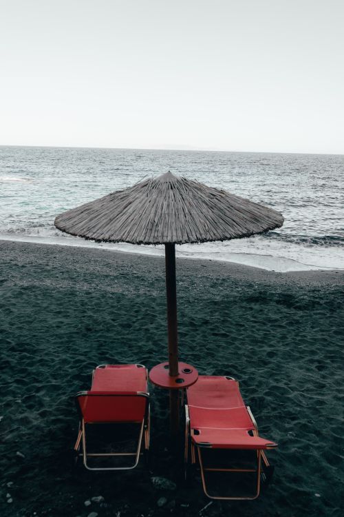 Free Relaxing view of red beach chairs under an umbrella facing the ocean in Mononaftis, Greece. Stock Photo