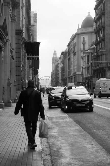 Black and white image of a man walking on a Barcelona street with cars and historic architecture.