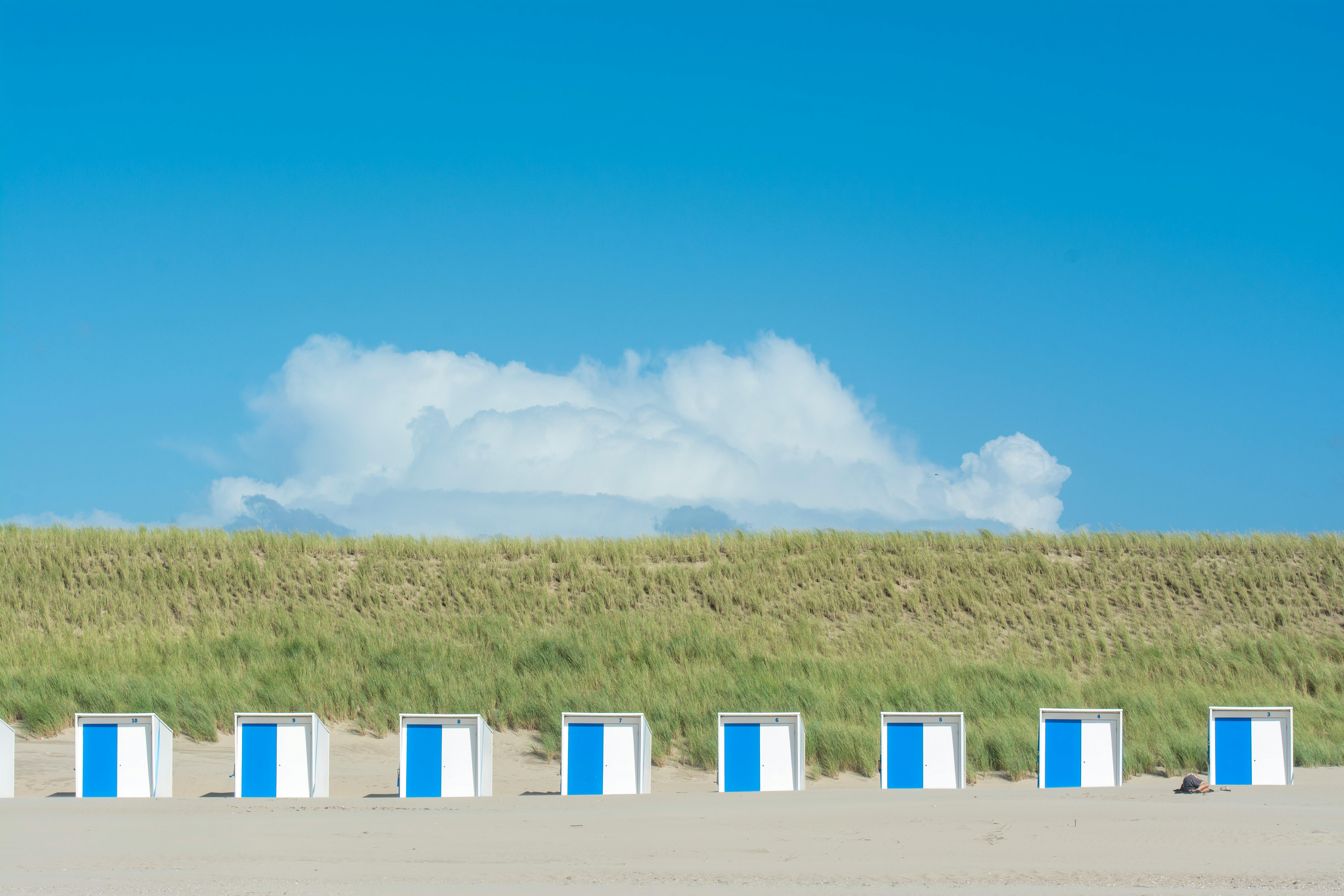 Line of blue and white beach cabins on sandy shore with green dunes and clear blue sky.