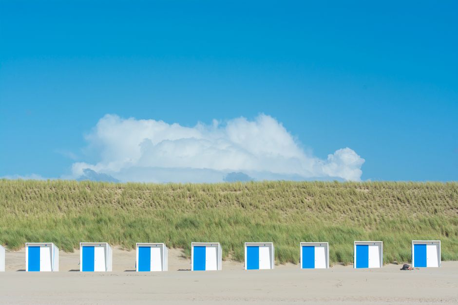 Line of blue and white beach cabins on sandy shore with green dunes and clear blue sky.