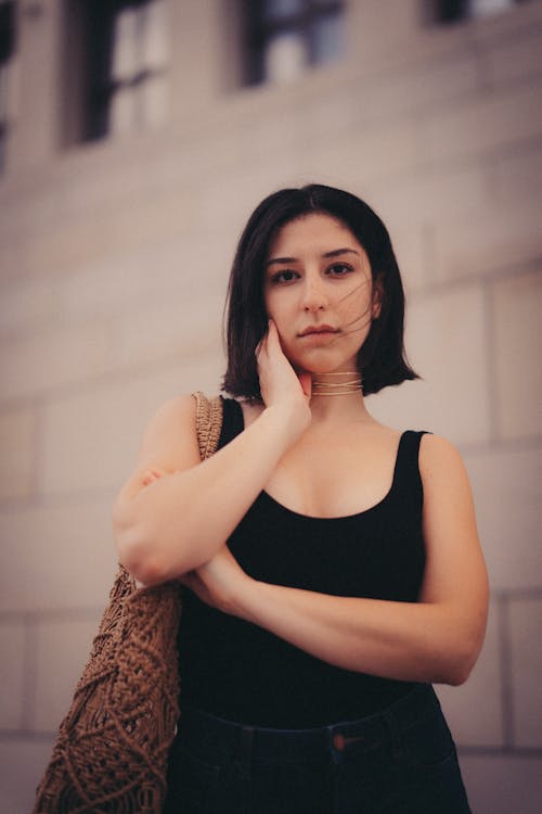 Young Woman in a Black Tank Top and Jeans Standing on the Background of a Building in City 