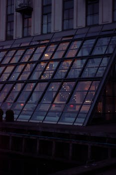 A modern building with a glass facade reflecting warm evening lights at dusk.