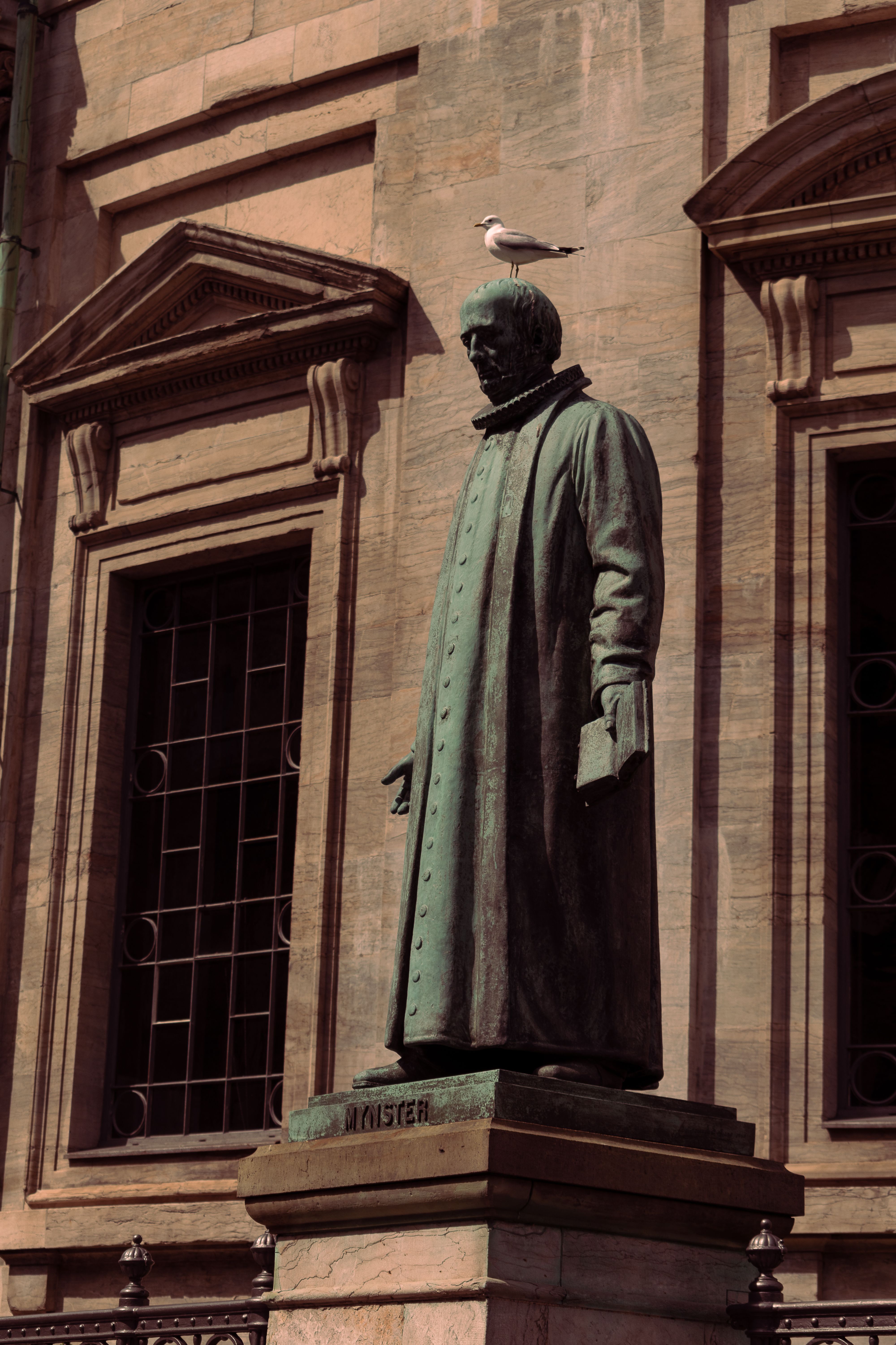 Bronze statue of a figure with a bird perched on its head, against historic architecture.