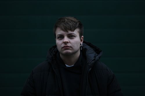 Free A contemplative young man in a dark jacket stands against a dark background. Stock Photo