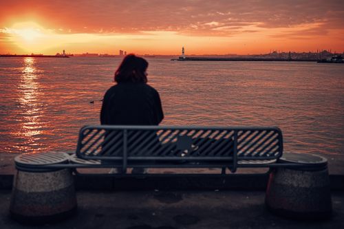 Free A woman sits on a bench by the sea, enjoying a serene sunset view. Stock Photo