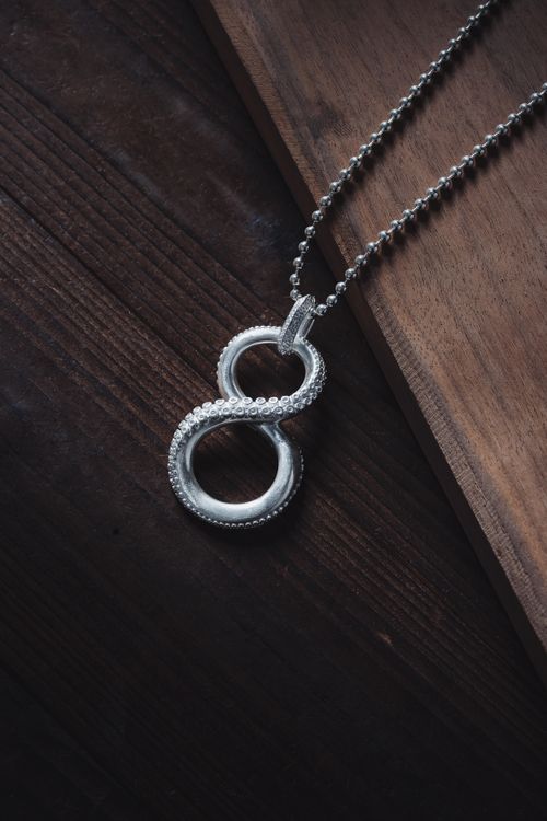 Free A close-up photo of a shiny infinity symbol necklace with metallic finish resting on a textured wooden surface. Stock Photo