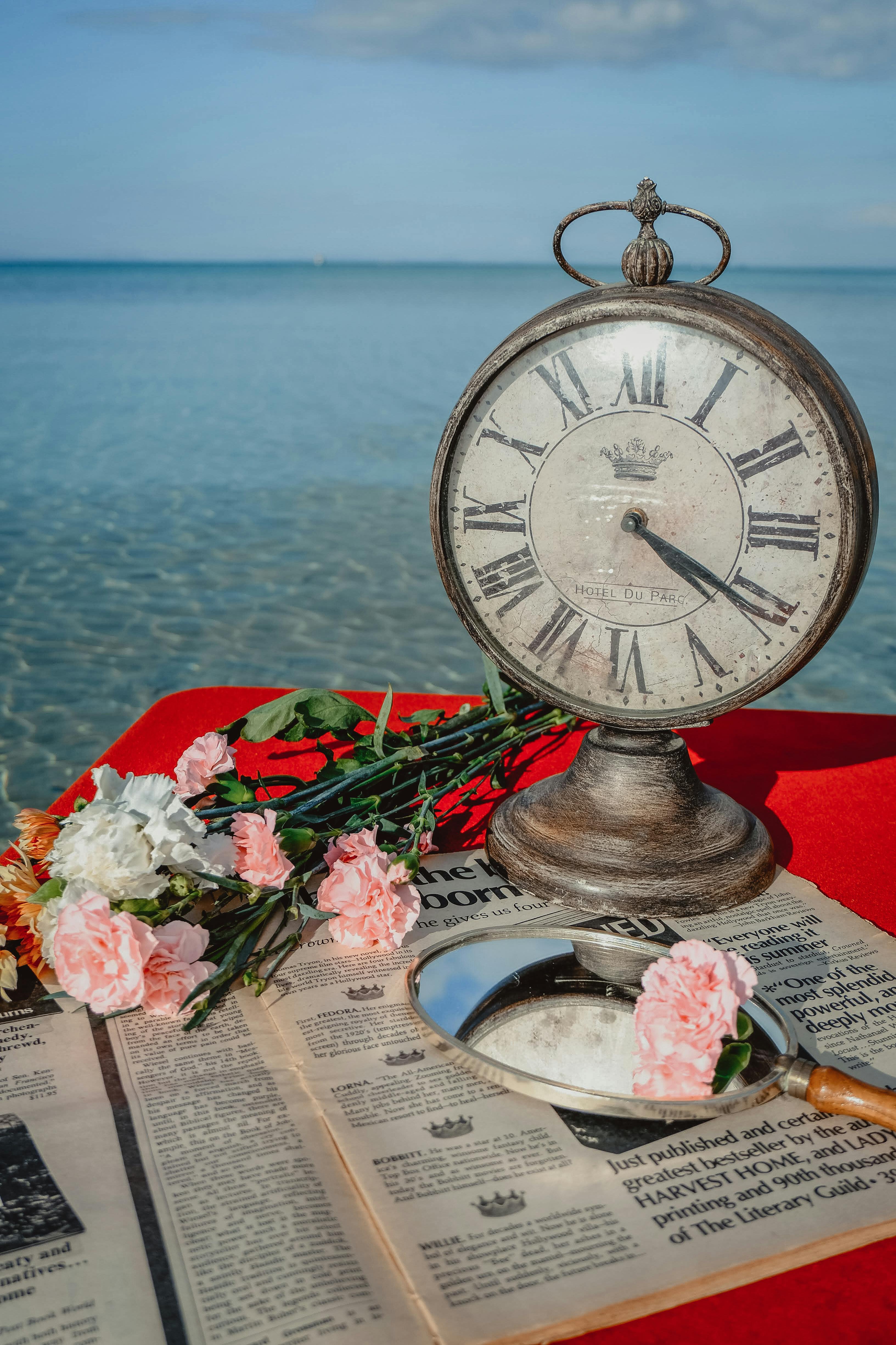 An antique clock and flowers on newspapers by the sea, evoking nostalgia and elegance.