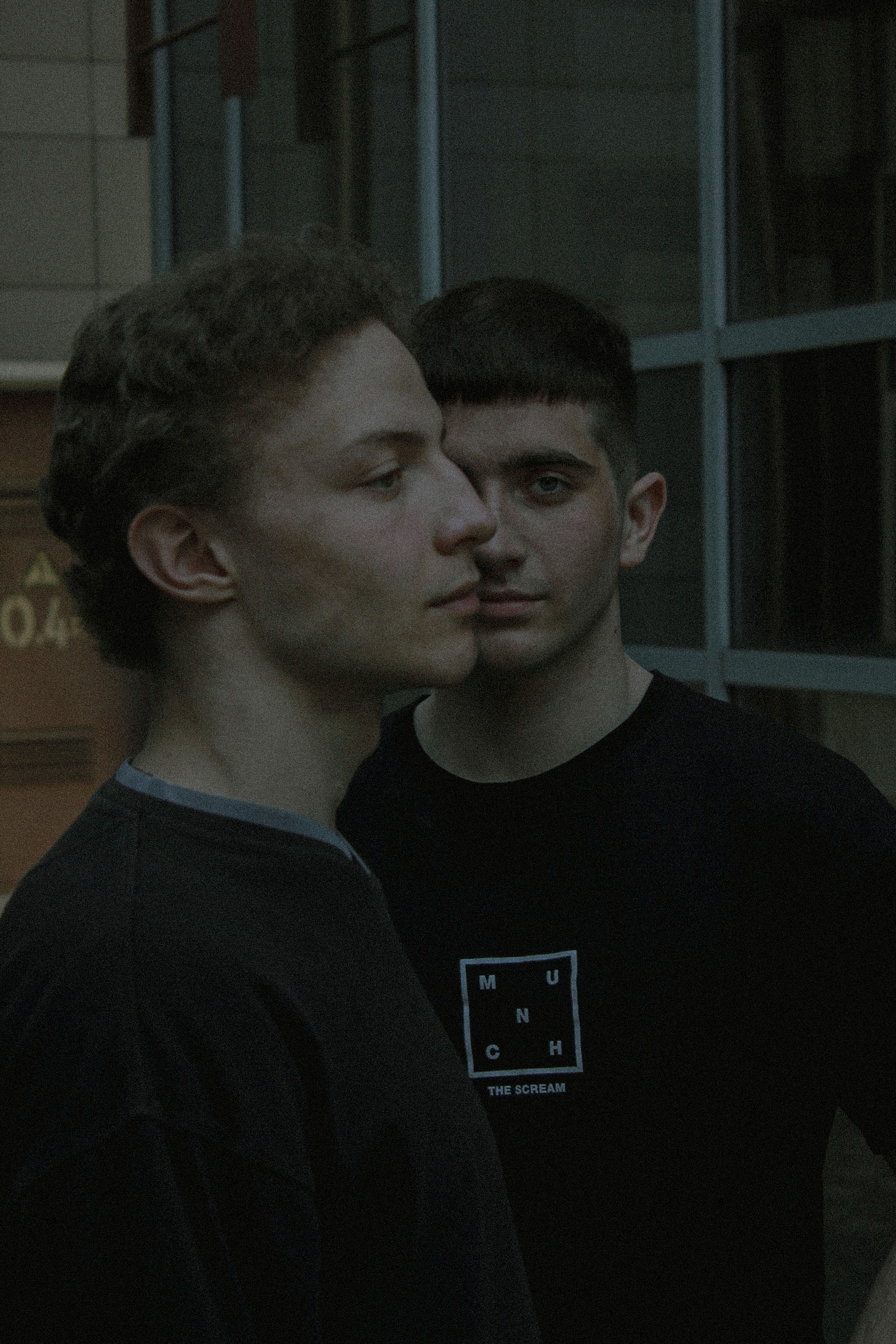 Artistic portrait of two young men with different haircuts posing near modern building windows.