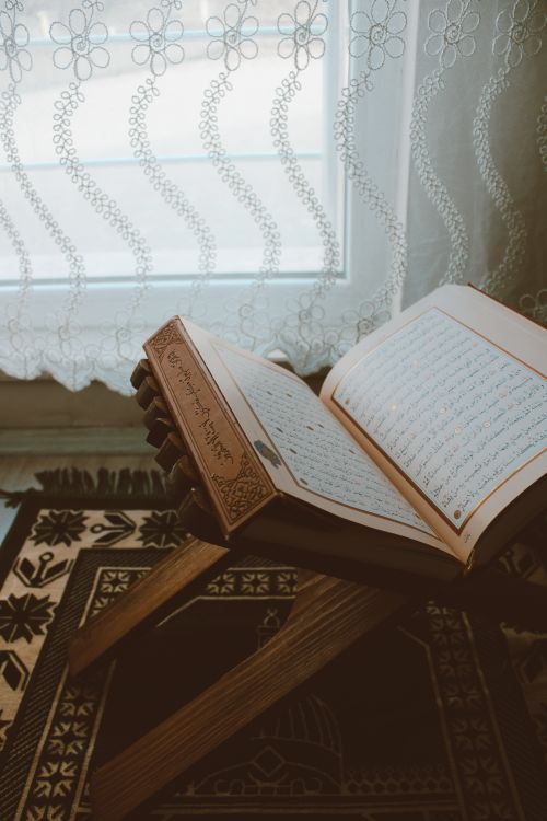 Free An open Quran rests on a wooden stand by a window, softly lit by natural light. Stock Photo