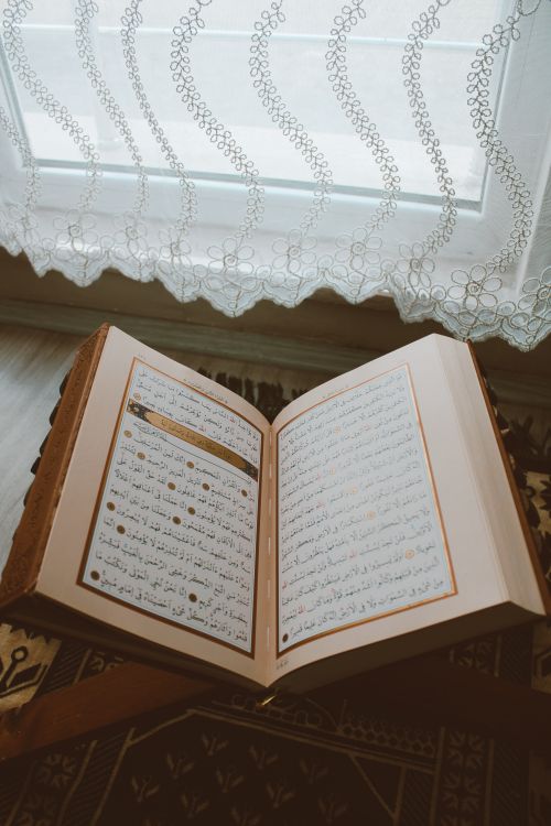 Free An open Quran on a wooden stand near a window with lace curtains. Stock Photo