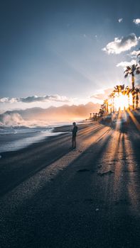 Sunset over Antalya beach with a silhouetted person walking amid ocean waves and palm trees.