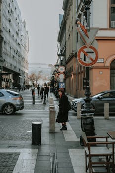 City street view in Budapest featuring pedestrians, parked cars, and historical architecture on a cloudy day.