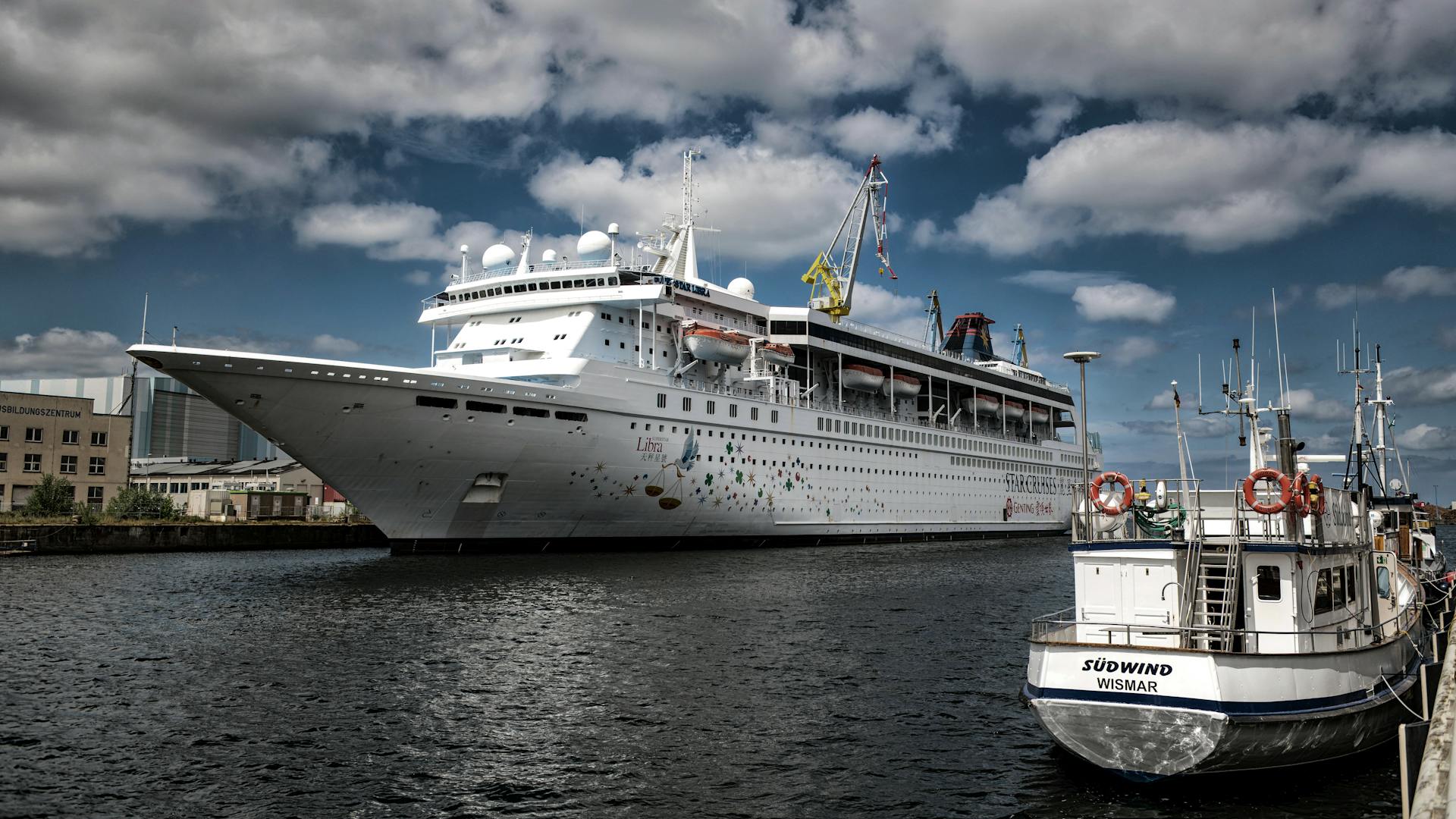 A large cruise ship and small boat docked in Wismar harbor, Germany, under a cloudy sky.