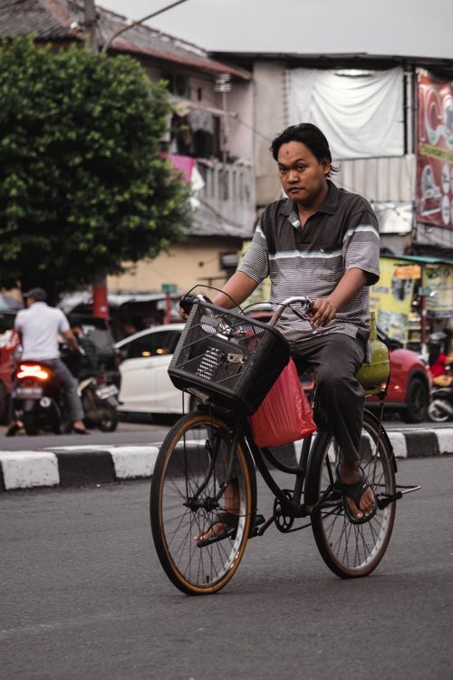 Free Man cycling through bustling urban street with parked cars and shops in the background. Stock Photo