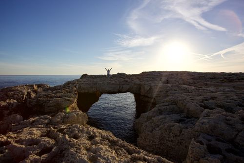 Free A person stands triumphantly on a natural rock arch overlooking the sea at sunrise. Stock Photo