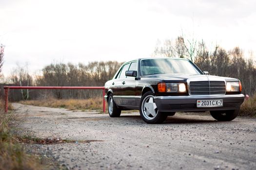 Classic black Mercedes Benz parked on a deserted road with autumn trees in the background, under a cloudy sky.