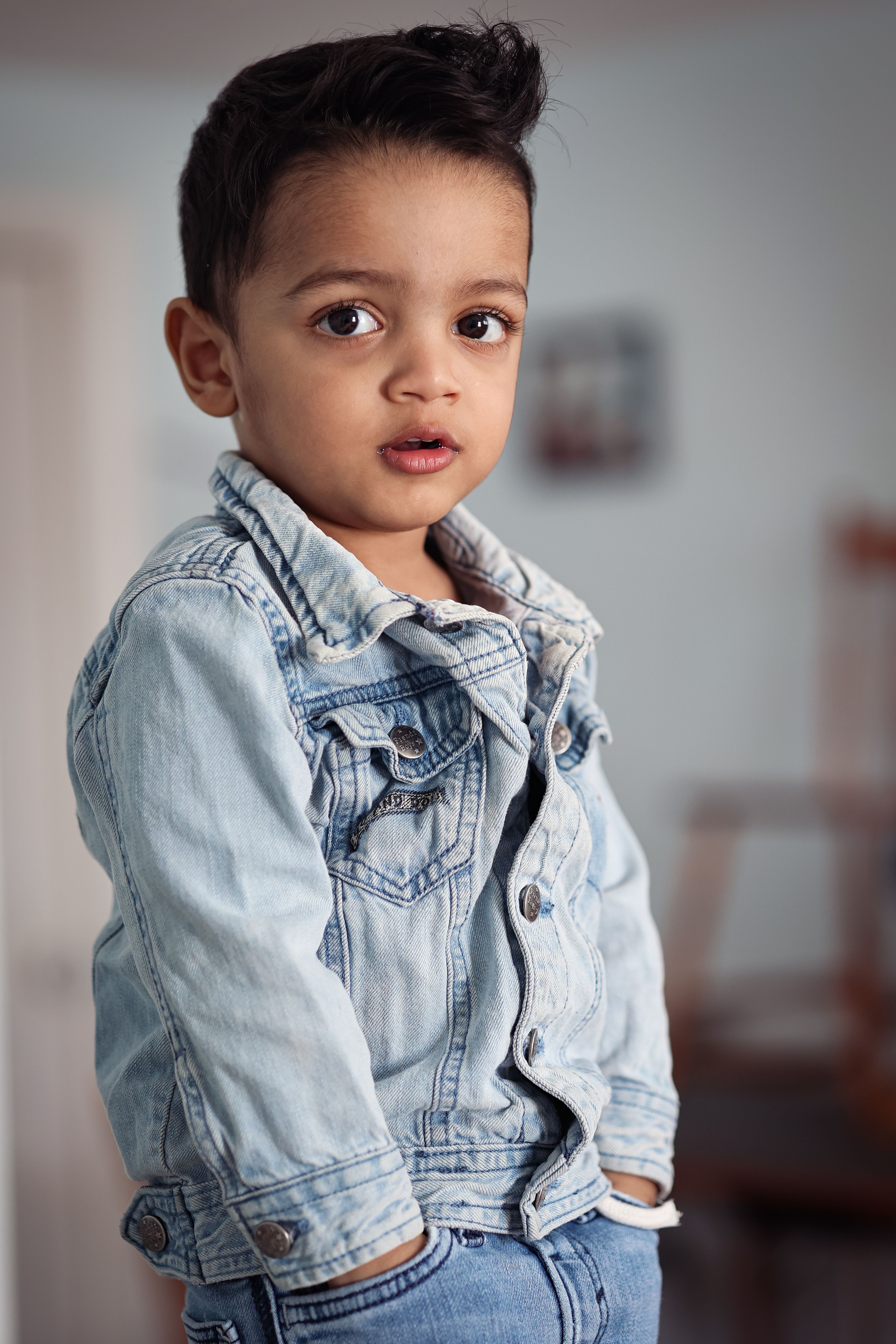 Portrait of a cute young boy in a denim jacket standing with hands in pockets indoors.