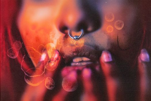 Close-up image of a face with piercing and hands, featuring artistic overlays and warm tones.