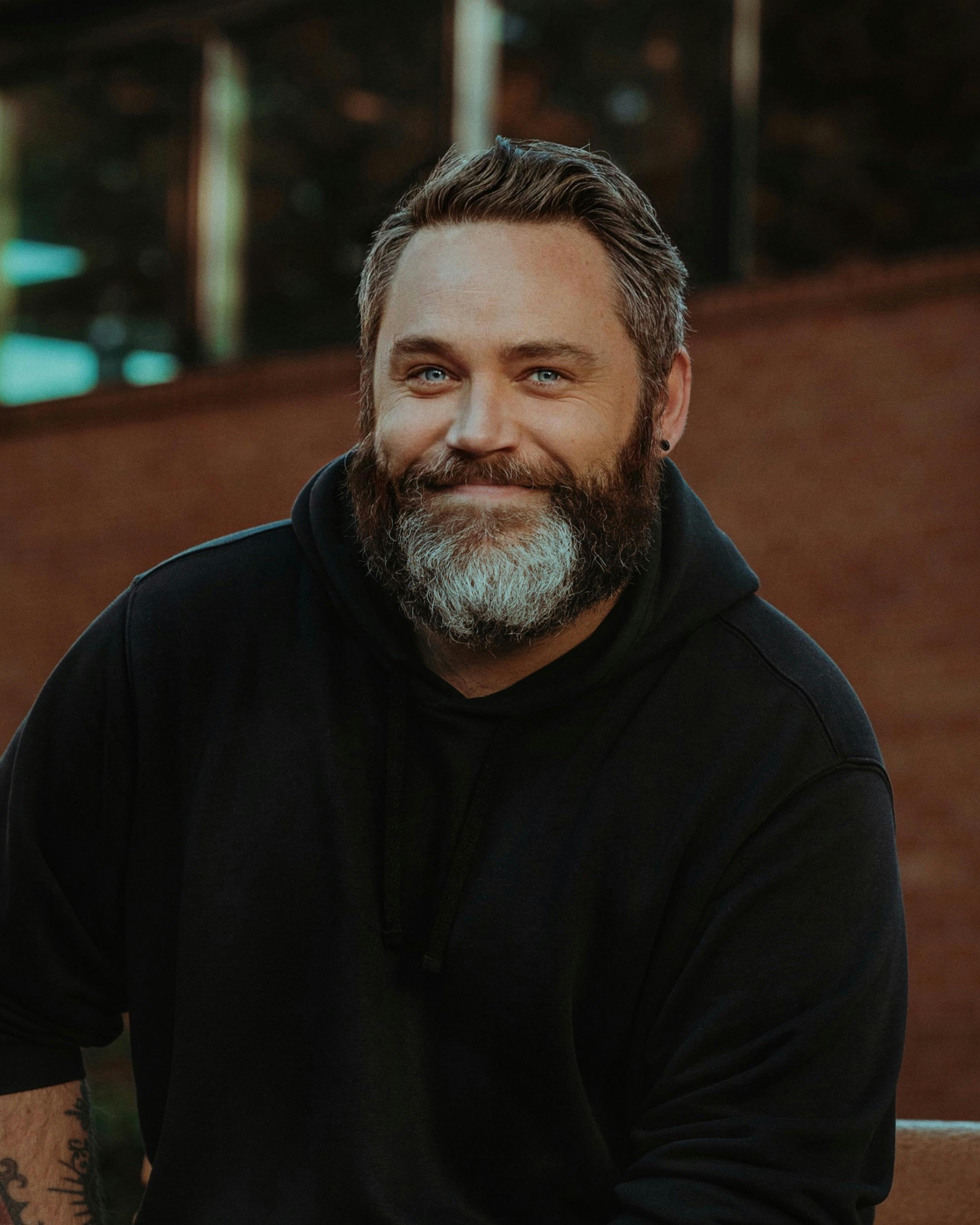 Portrait of a smiling bearded man in a black hoodie outdoors, creating a warm and approachable vibe.
