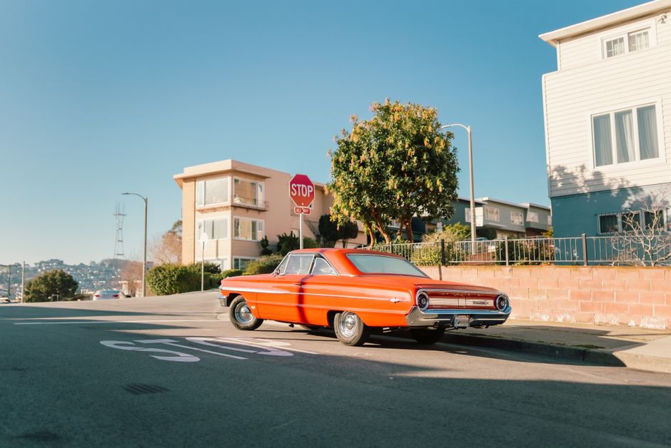 A vibrant vintage red car parked at a sunlit urban crossroads with clear blue skies, stop sign in view.