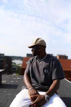 Man in cap on rooftop with city background, daytime outdoor shot.