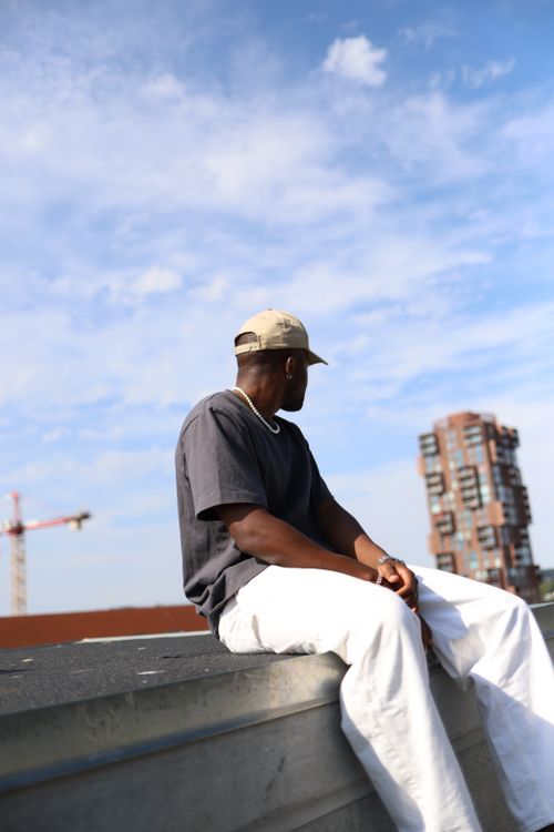 Free A man in a cap and t-shirt sits on a rooftop overlooking a cityscape, under a blue sky with clouds. Stock Photo