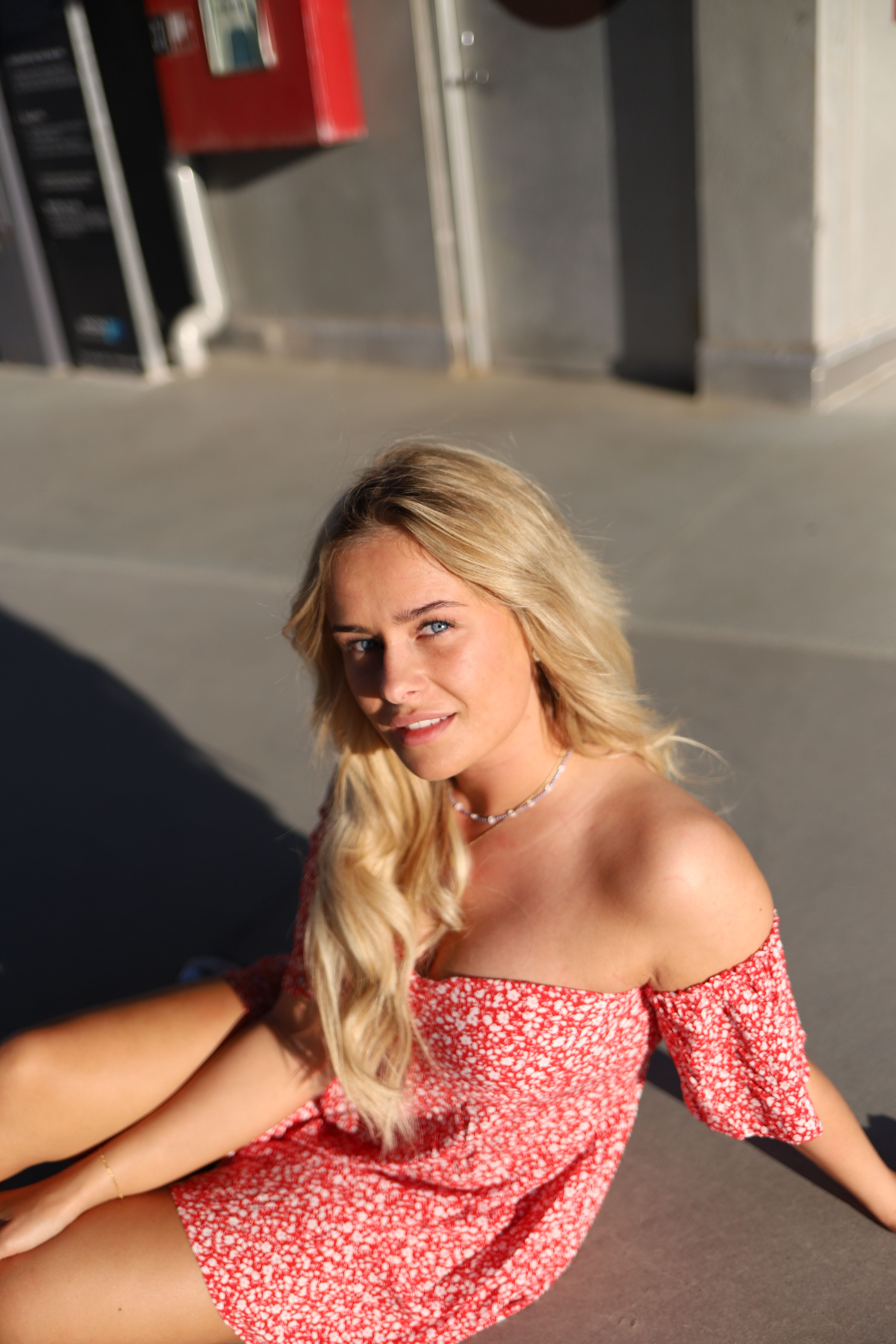 Woman with blonde hair sitting on pavement in a sunlit setting, wearing a red floral dress.