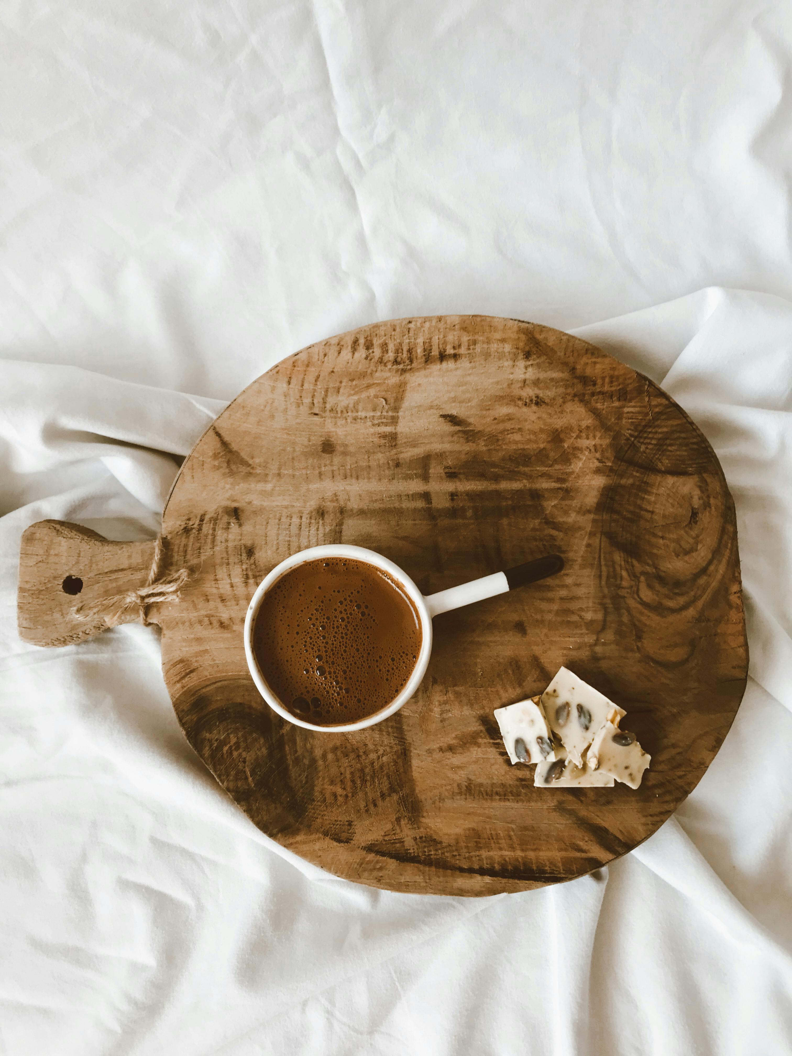 Rustic wooden cutting board with coffee cup and cheese placed on white fabric backdrop.