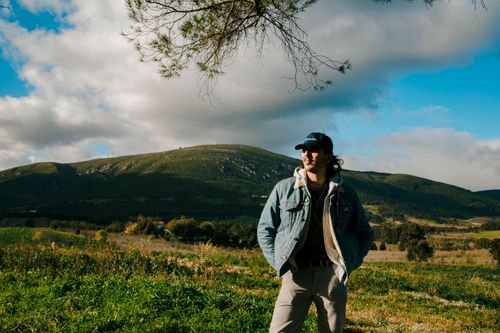 Free A man wearing a cap and jacket stands outdoors against a panoramic hill view. Stock Photo