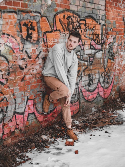 Male Model Wearing a Gray Sweatshirt Standing on One Leg in Front of a Brick Wall