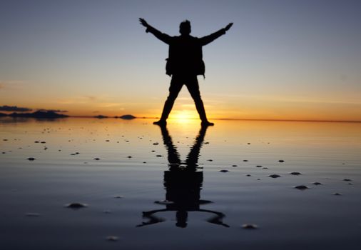 Silhouette of a person with arms raised at sunset, reflected on a calm coastal shore.