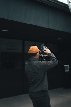 Man in orange beanie filming city scene outdoors, showcasing urban video production.