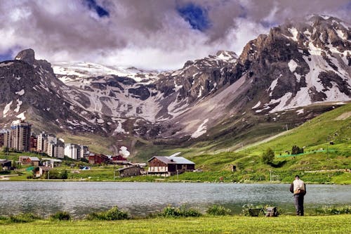 Man in Beige and Black Jacket Standing on Green Grass Lawn in Front of Black and Gray Mountain