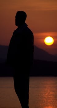 A silhouette of a man standing by the sea against a vibrant sunset sky.