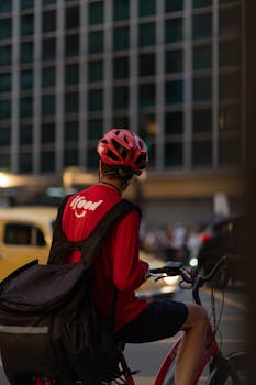 Back view of a food delivery cyclist in urban setting wearing a helmet and carrying a delivery bag.