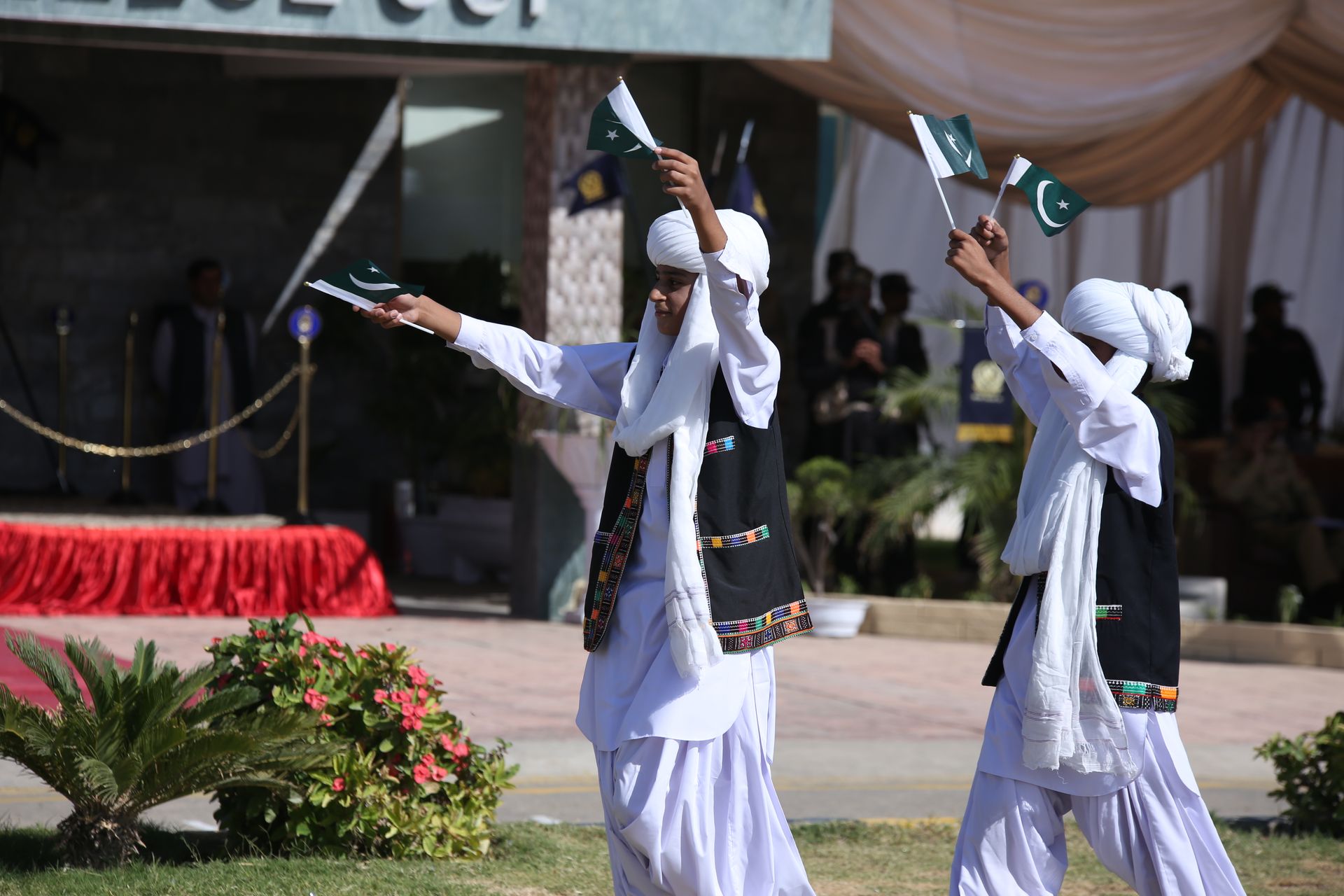 Balochi boys in traditional attire waving Pakistani flags during an outdoor celebration.