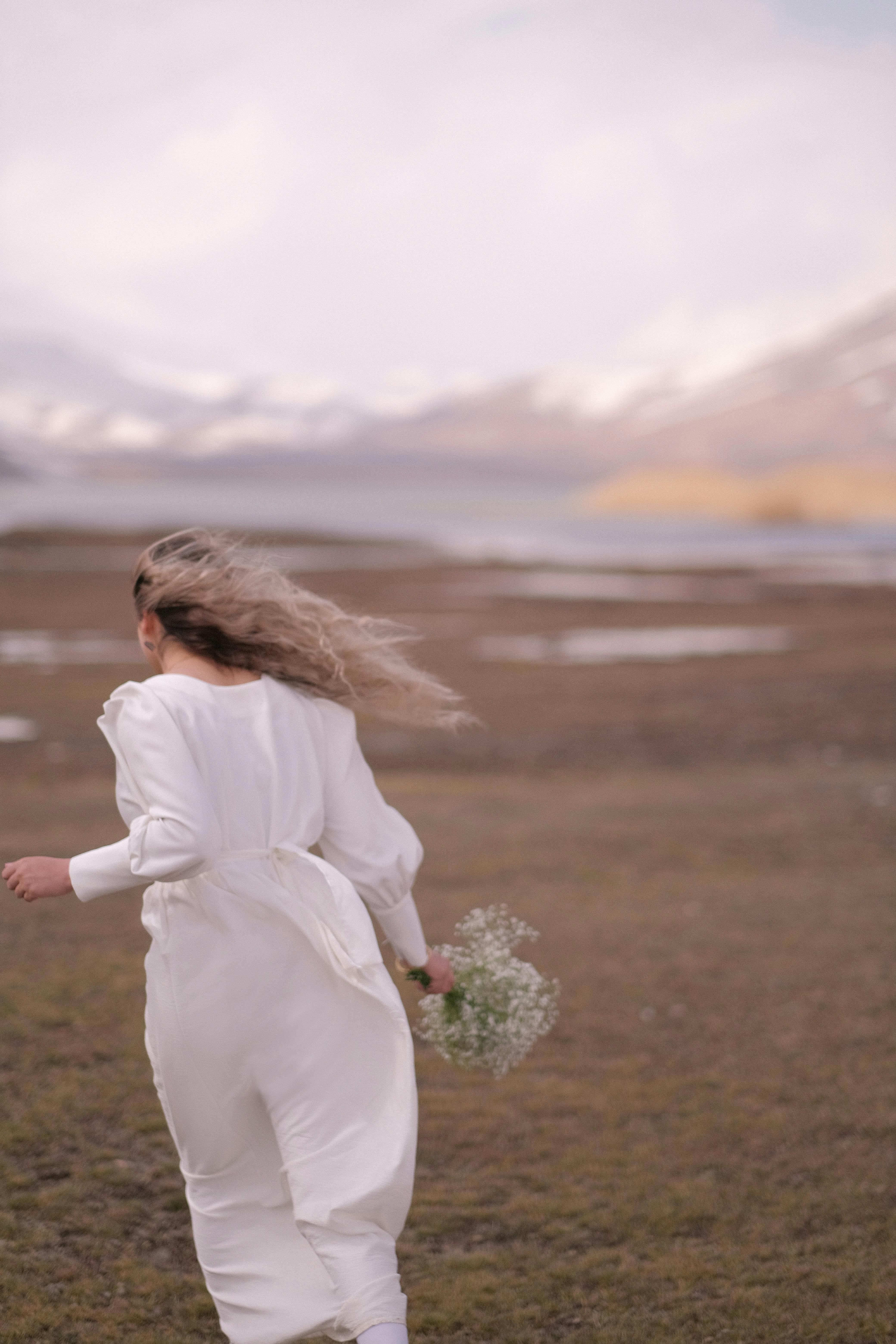 Woman in a flowing white dress runs through a scenic landscape holding a bouquet of flowers.
