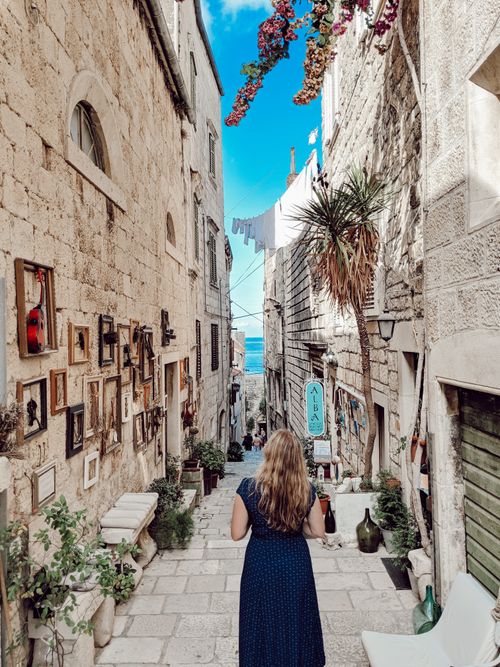 Free Back view of a woman walking in Dubrovnik's charming stone alley with artworks and plants. Stock Photo